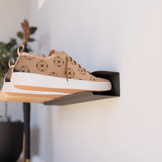 Wall-mounted shoe rack in black, holding a pair of brown patterned sneakers, installed on a white wall next to a potted plant with a wooden floor in the background.