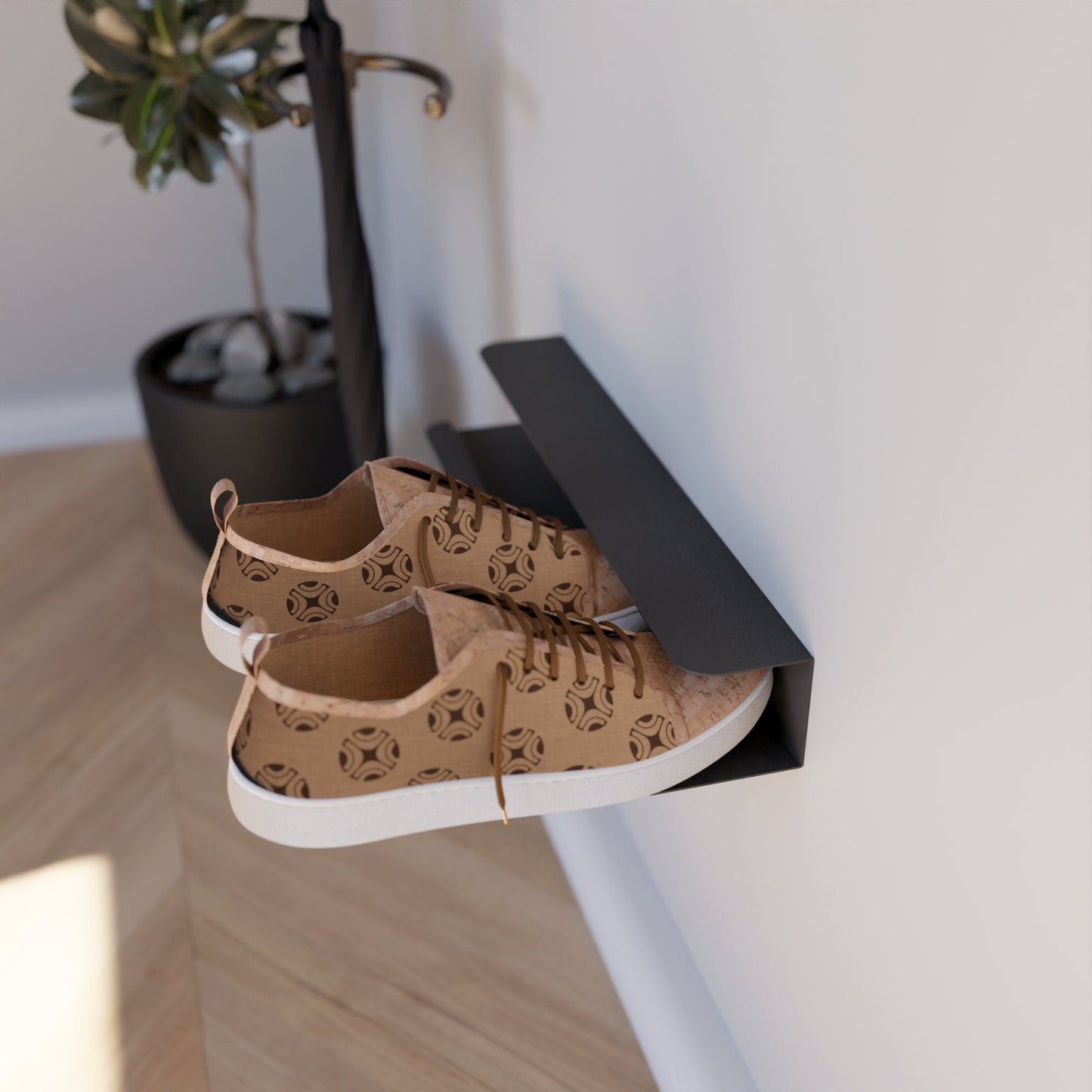 Wall-mounted shoe rack in black, holding a pair of brown patterned sneakers, installed on a white wall next to a potted plant with a wooden floor in the background.